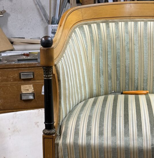 Poltrona antica stile Impero con vecchio rivestimento usurato a righe verticali verdi e panna in un laboratorio di tappezzeria.