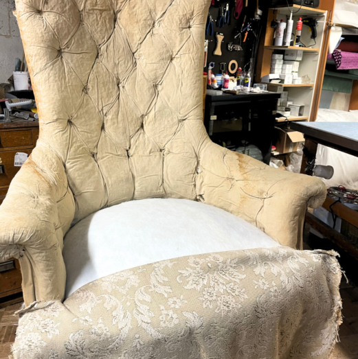Poltrona vintage capitonné beige usurata all'interno di un laboratorio di tappezzeria prima del rifacimento.